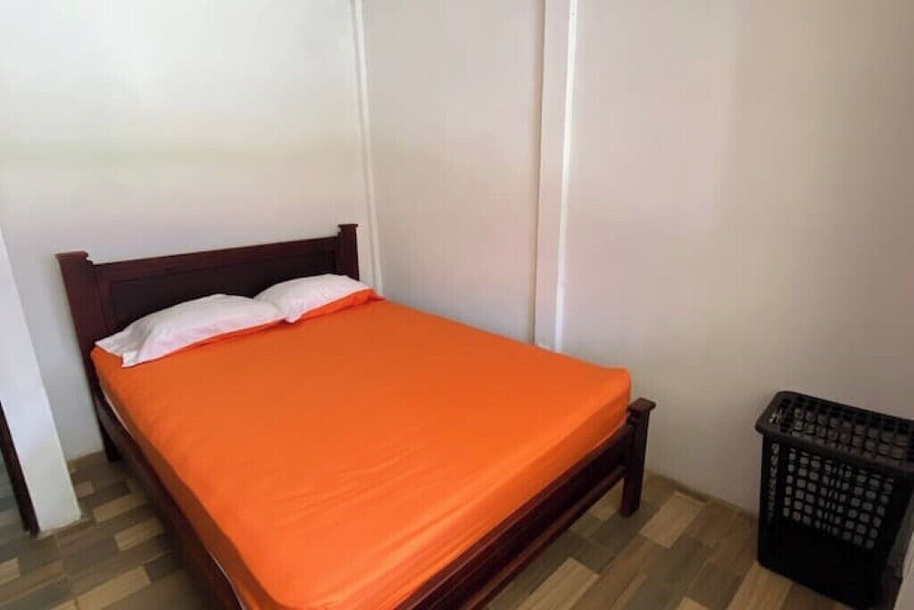 A single bed with an orange bedspread and white pillows in a room with wooden flooring and white walls.