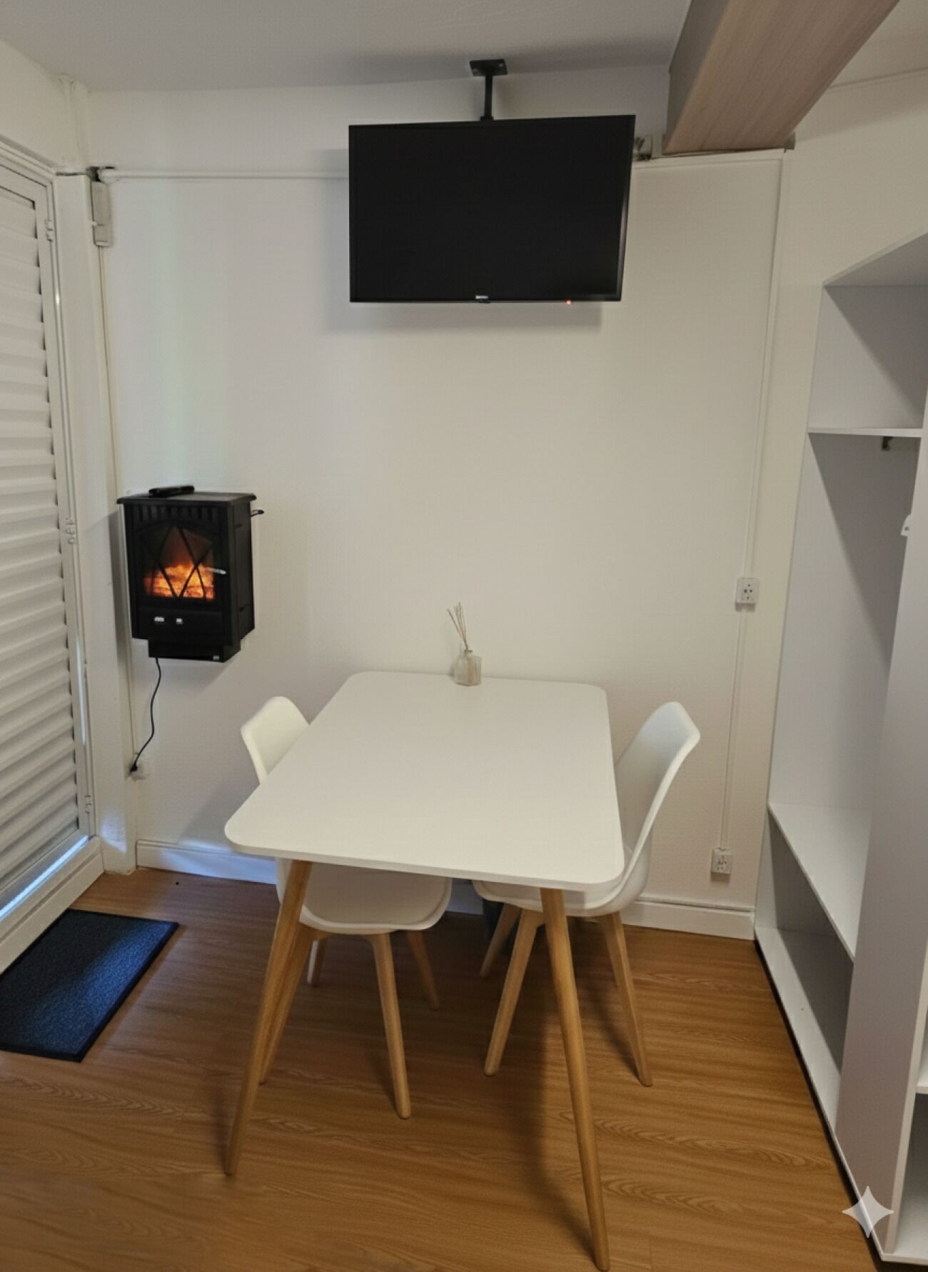 Una habitación pequeña con una mesa blanca, dos sillas, un televisor de pantalla plana montado en la pared y un estufa de leña.
