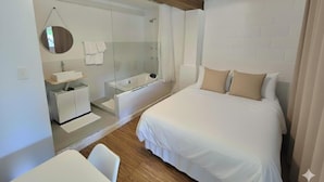 Una habitación de hotel moderna con una cama grande, un baño separado con ducha de vidrio, un escritorio pequeño y un espejo redondo.