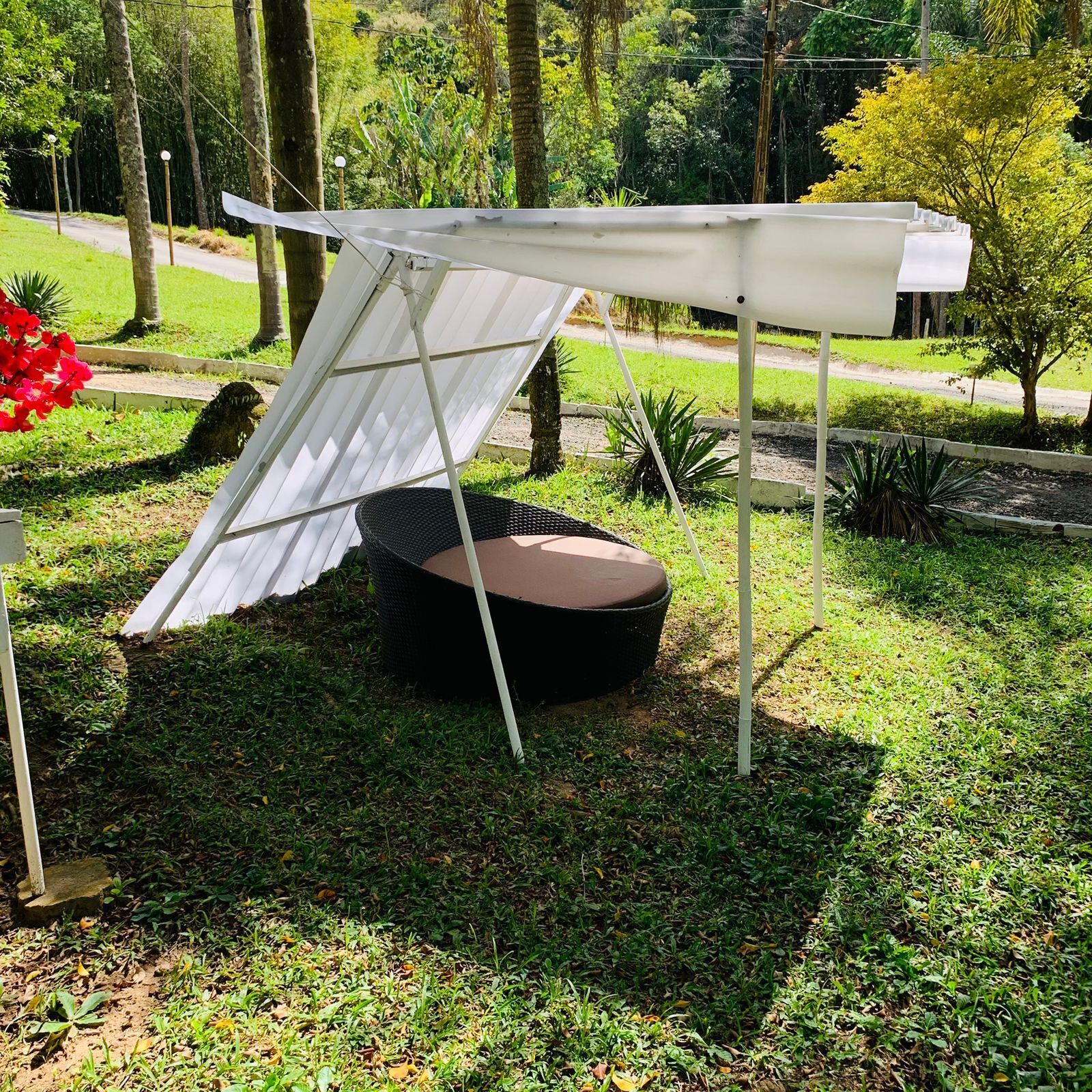 Un espacio al aire libre con toldo blanco, una mesa redonda de mimbre negro y un césped con árboles.