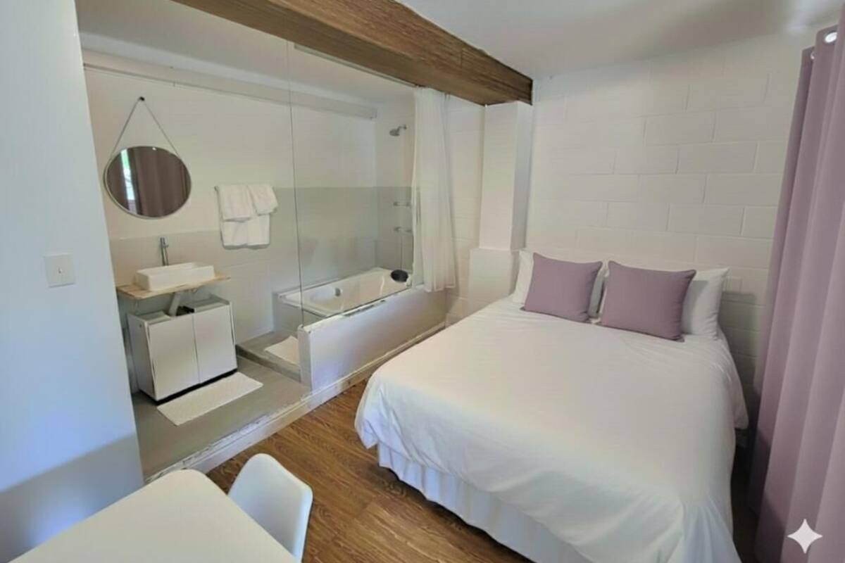 A modern bedroom with a large bed, a bedside table, a mirror, a shower area, and a small seating area.