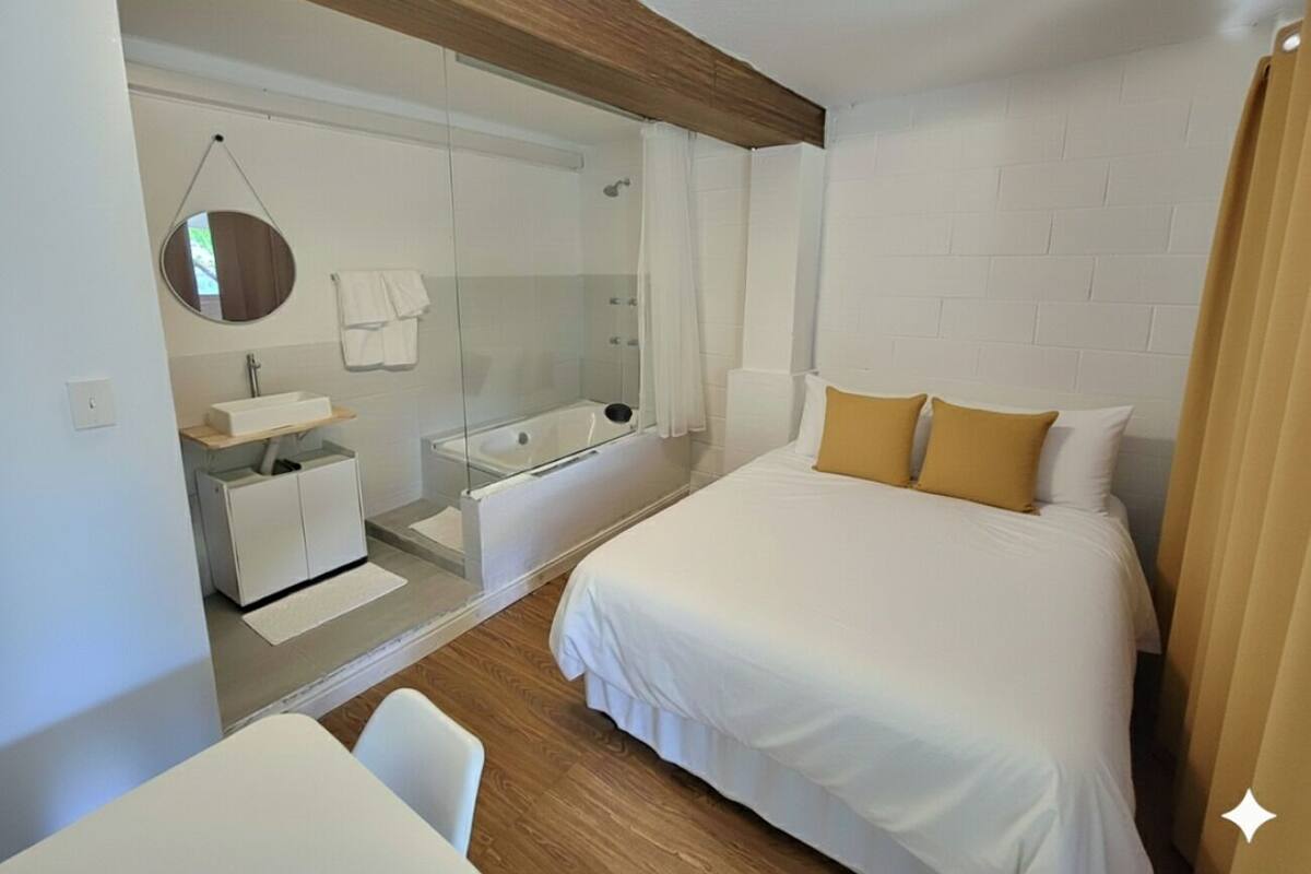 A modern hotel room with a large bed, a glass-enclosed shower area, a small white desk, and a round mirror on the wall.