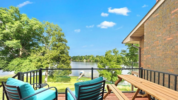 Una terraza de madera con dos sillas y una mesa, con vista a un lago que incluye un bote, rodeada de árboles.
