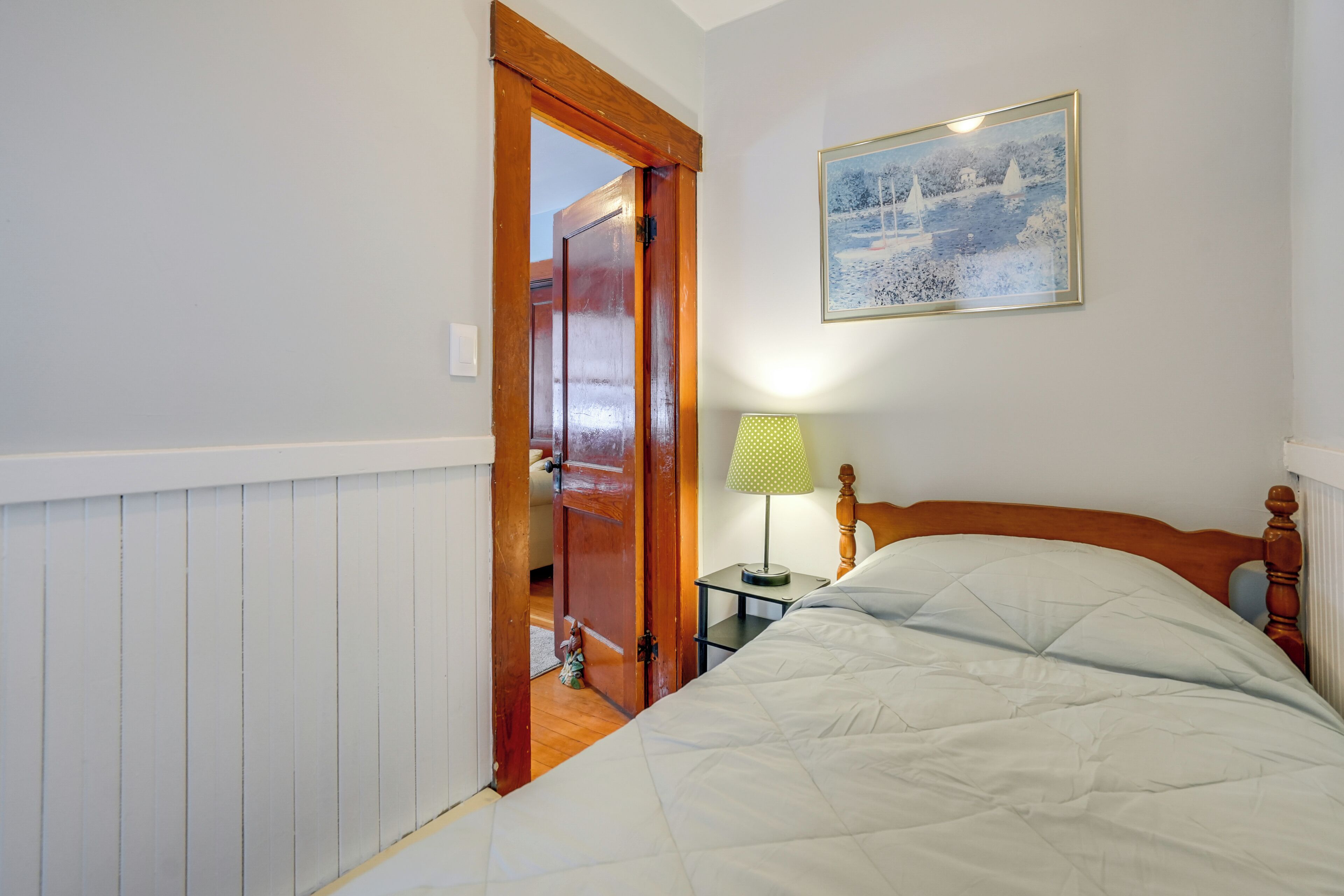A bedroom with a wooden door, a bed with a quilted blanket, a nightstand with a lamp, and a framed picture on the wall.