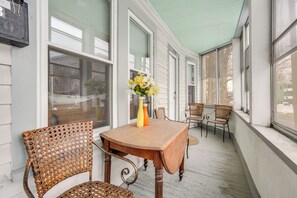 Une véranda avec une table en bois et des chaises en osier, un vase rempli de fleurs et de grandes fenêtres.