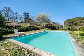 A rectangular swimming pool with a tiled deck, surrounded by greenery and trees.