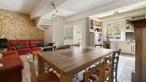 A spacious living area with a stone wall, a red leather sofa, a wooden dining table, and a kitchen with white cabinets and a stone floor. - Gîte de Marlas - Ariège (Rieucros)