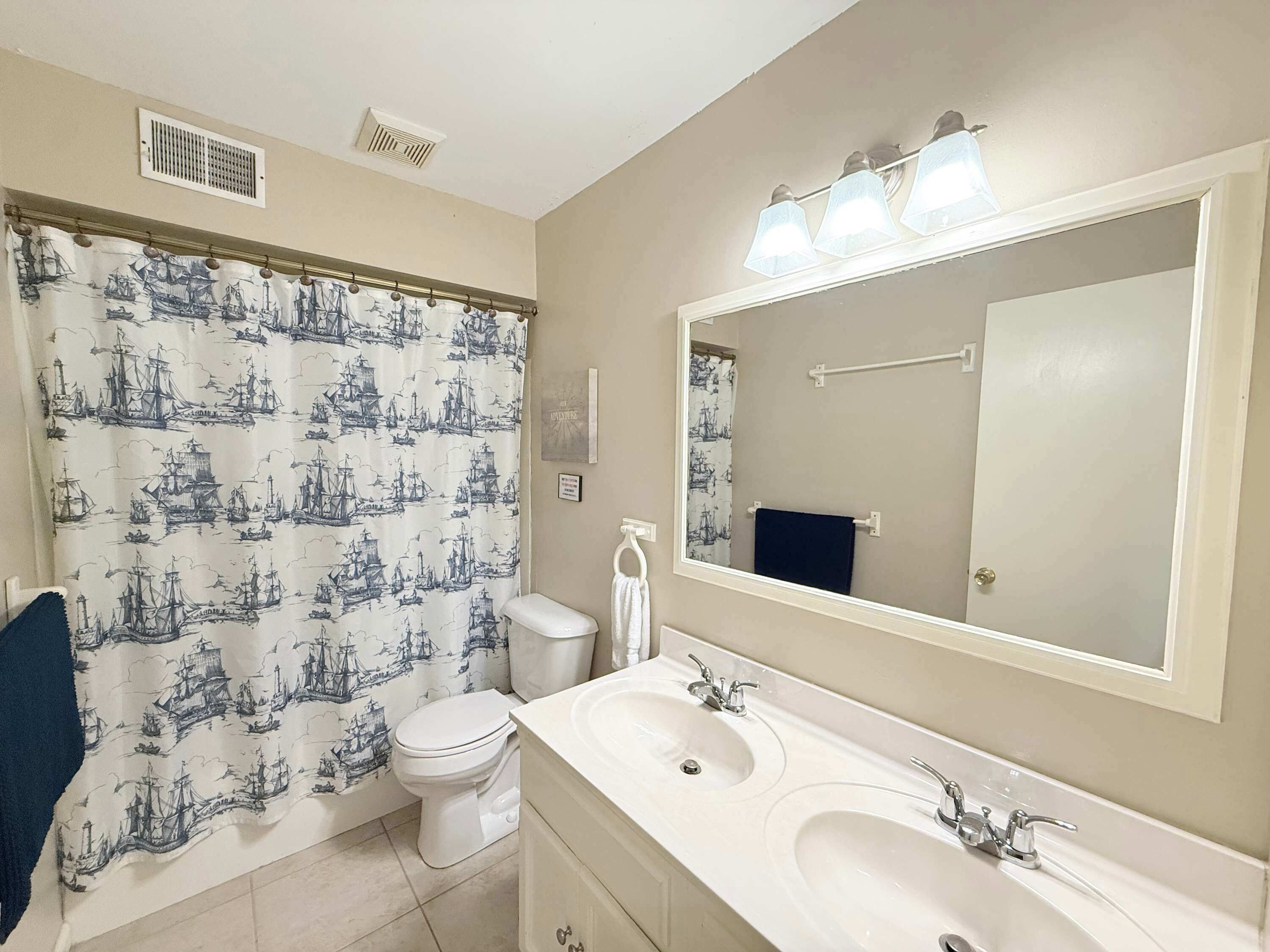 A bathroom with a ship-themed shower curtain, a toilet, a sink with two basins, and a large mirror.