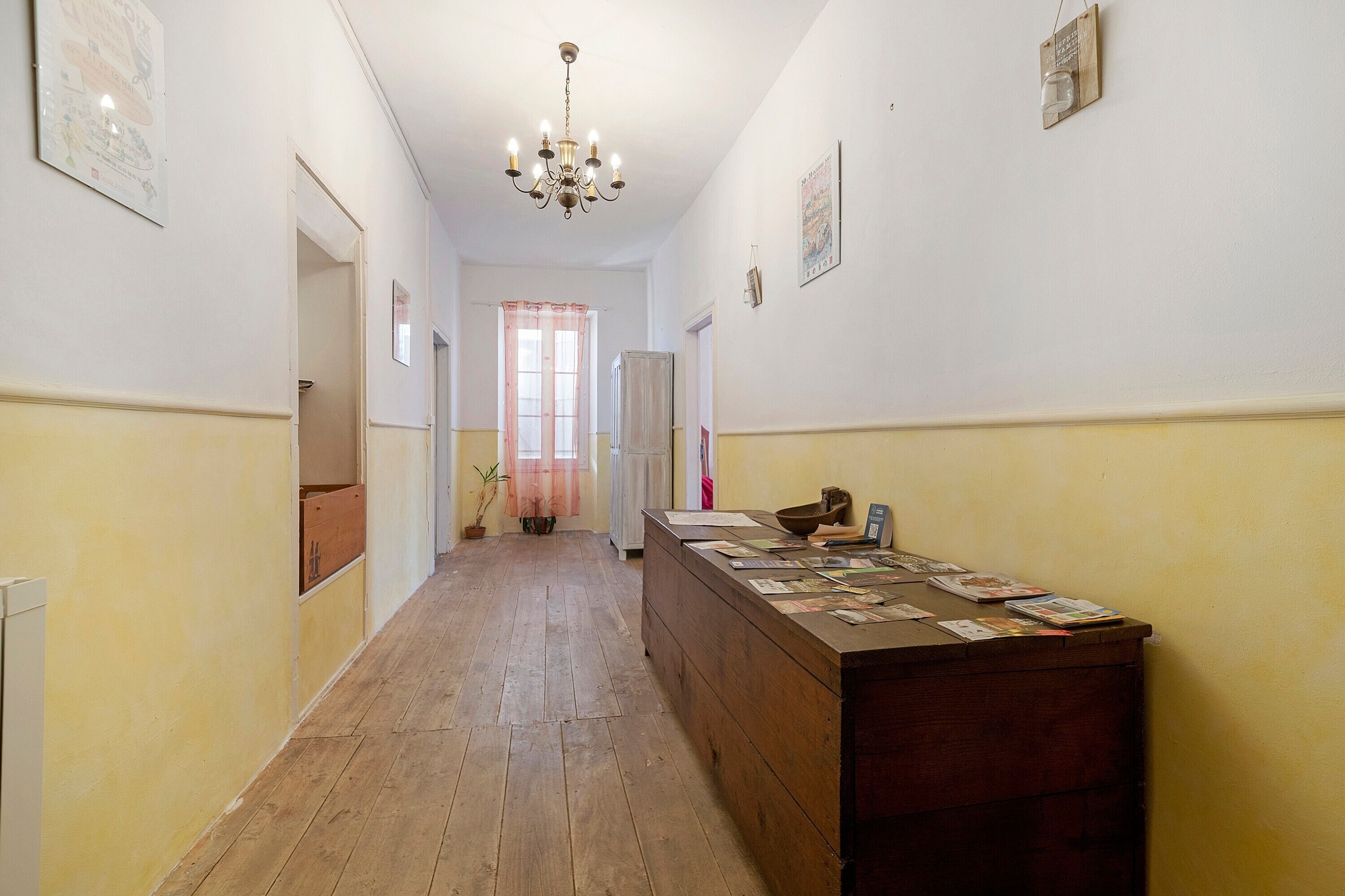A hallway with a wooden floor, a desk with papers, a chandelier, and a window with curtains.