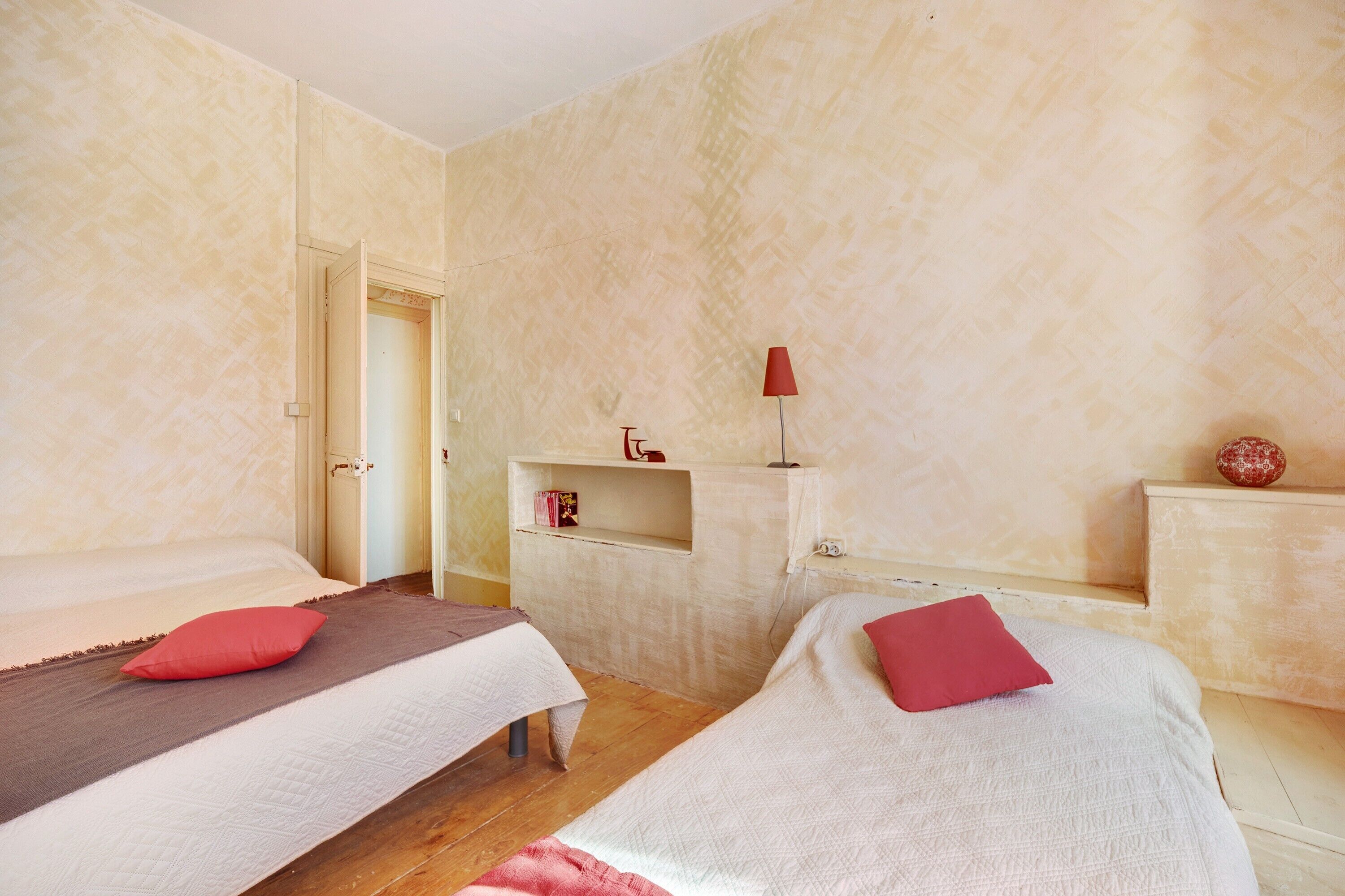 Une chambre avec deux lits, une étagère sur laquelle se trouve une lampe rouge, et une porte qui donne sur une autre pièce.