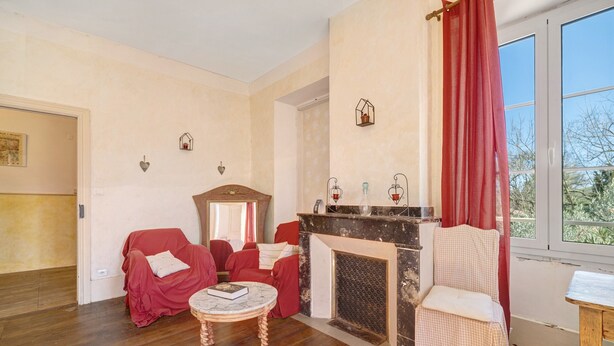 Une pièce avec une cheminée, un fauteuil rouge, une petite table avec un livre et une fenêtre avec des rideaux.