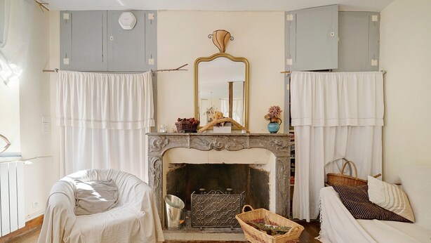 Une pièce avec une cheminée, un miroir, un canapé, un fauteuil et une fenêtre avec des rideaux.