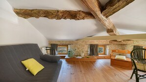 Une pièce de style loft avec des poutres apparentes, un canapé gris agrémenté d’un coussin jaune, un banc en bois et une chaise verte.