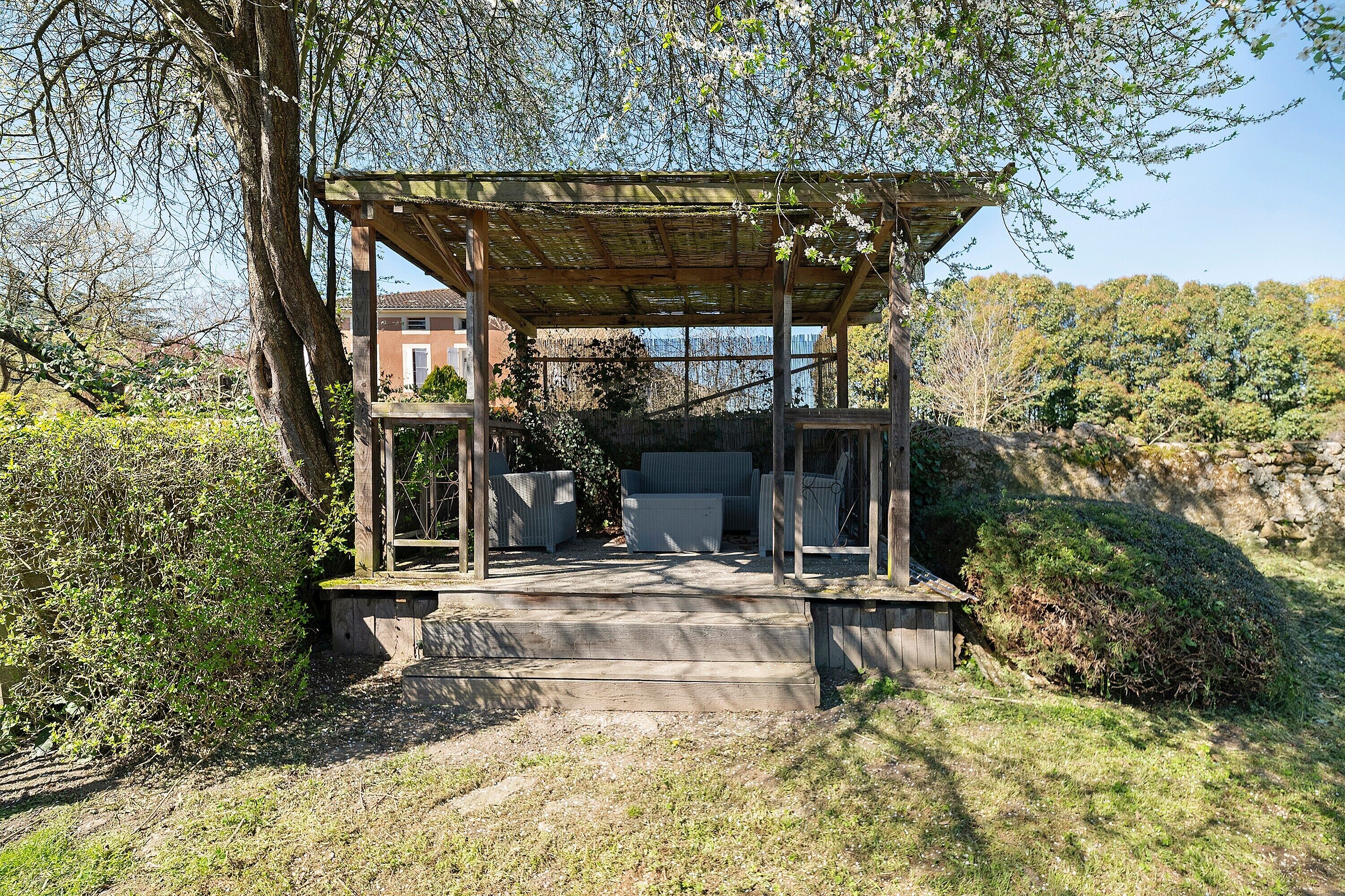 Een houten pergola met een wenteltrap erheen, omgeven door groen en een stenen muur.
