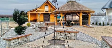 Une cabane en bois dotée d’un espace extérieur avec toit de chaume, d’un foyer en pierre et d’un jardin clôturé.