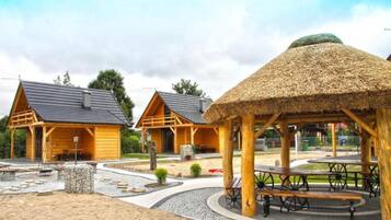 Une cabane en bois avec un toit de chaume, un espace pique-nique avec des bancs et un sentier en pierre.