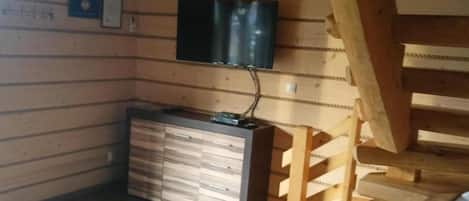 Una habitación con una escalera de madera, un televisor de pantalla plana montado en la pared y un mueble de madera con cajones.