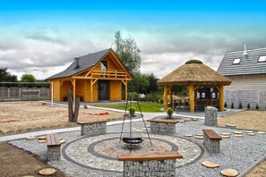 A wooden cabin with a thatched-roof gazebo, surrounded by a gravel area with benches and a circular stone feature.