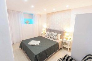 A hotel room with a bed, bedside lamps, a nightstand, a window with curtains, and a small plant on the nightstand. - Eidolion Essence - Cozy Guest House Mykonos (Ano Mera)