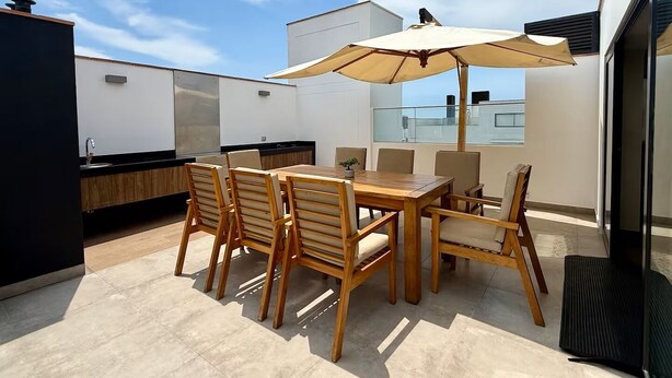 A rooftop patio with a wooden table and chairs, a large umbrella, and a modern kitchen area. - APT GRAÑA - MAGDALENA  (Lima)