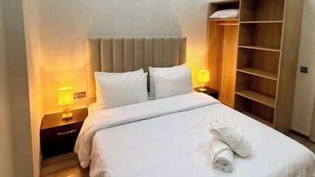 Un lit bien fait, avec du linge de lit blanc, une table de chevet en bois avec une lampe et une armoire intégrée en bois.