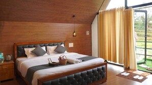 A bedroom with a wooden slatted ceiling, a large bed with a black upholstered headboard, and a view of a green landscape through a window with yellow curtains. - Airas Resort (Kodaikanal)
