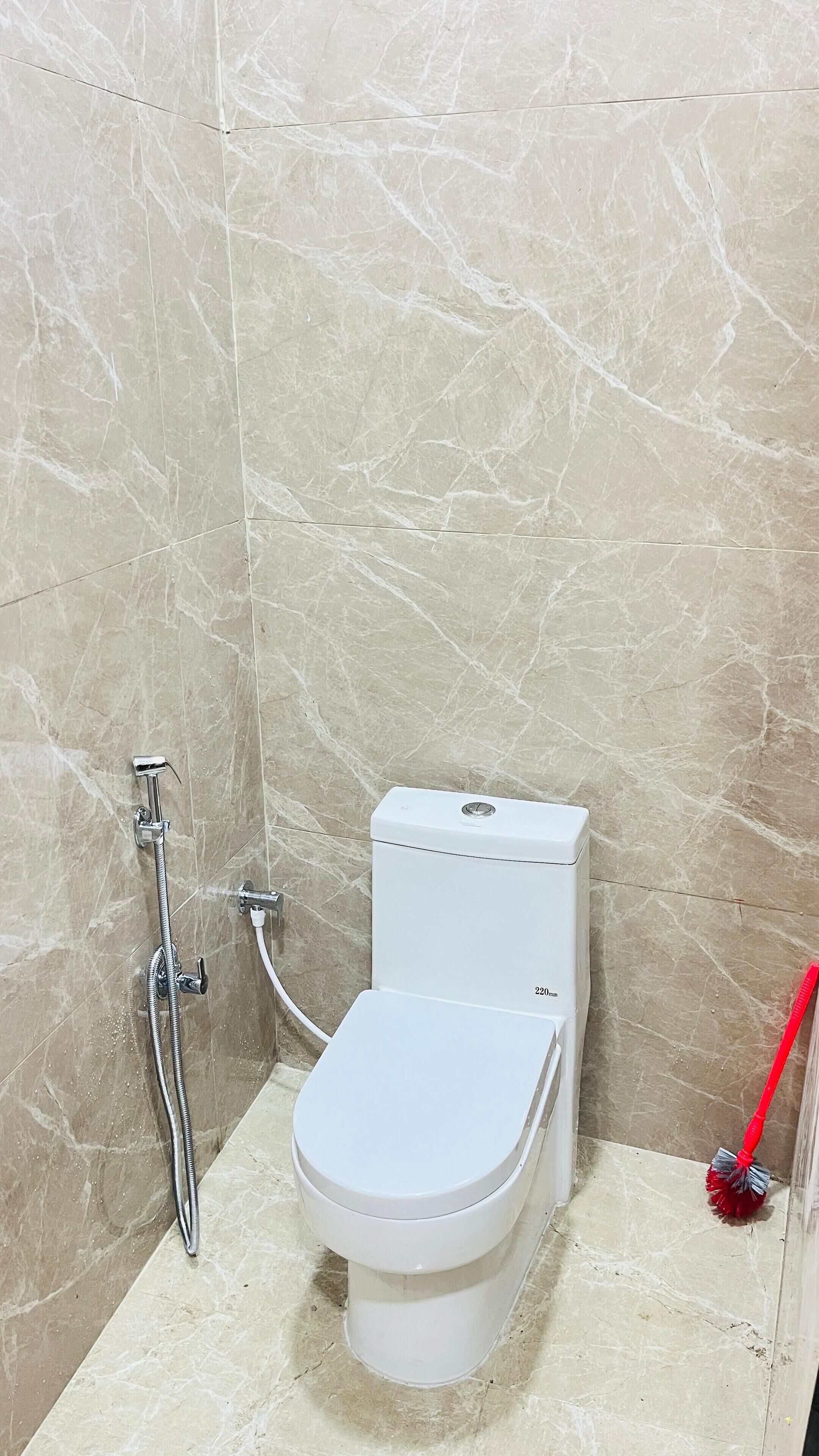 A small, tiled bathroom with a toilet, a showerhead, and a red cleaning brush.