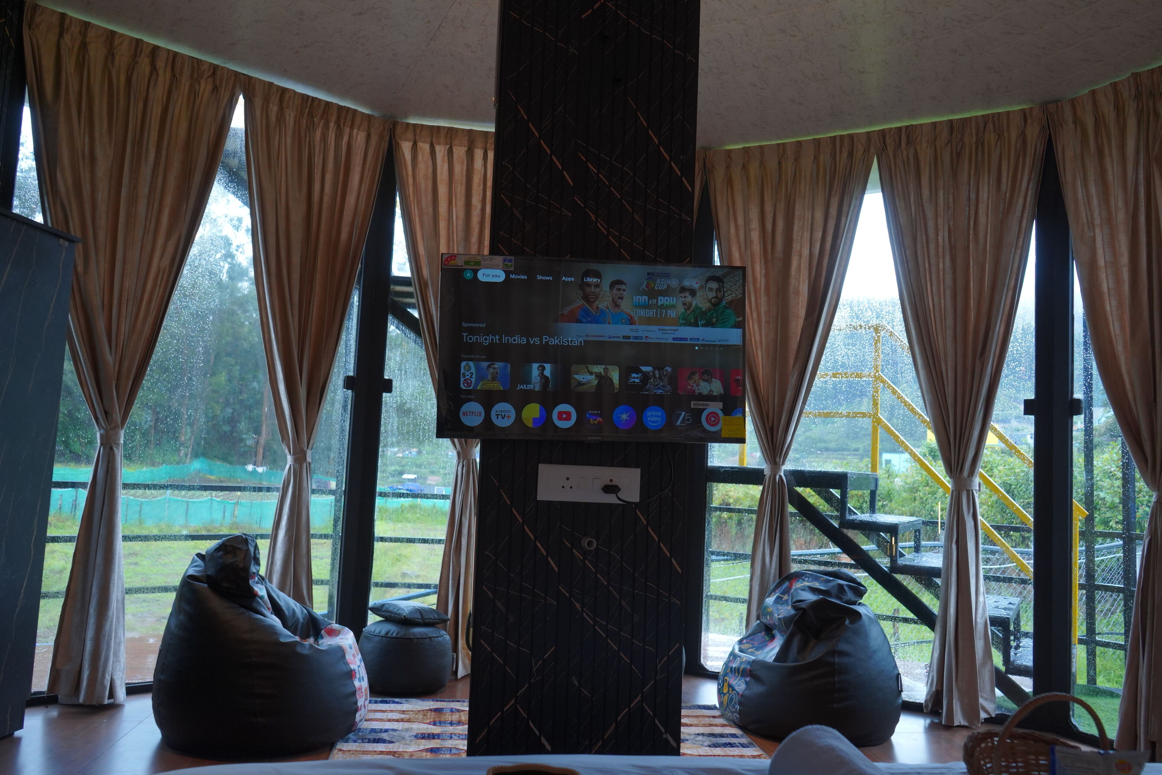 A room with a large TV screen displaying a sports event, bean bags, and a view of a green outdoor area through large windows.