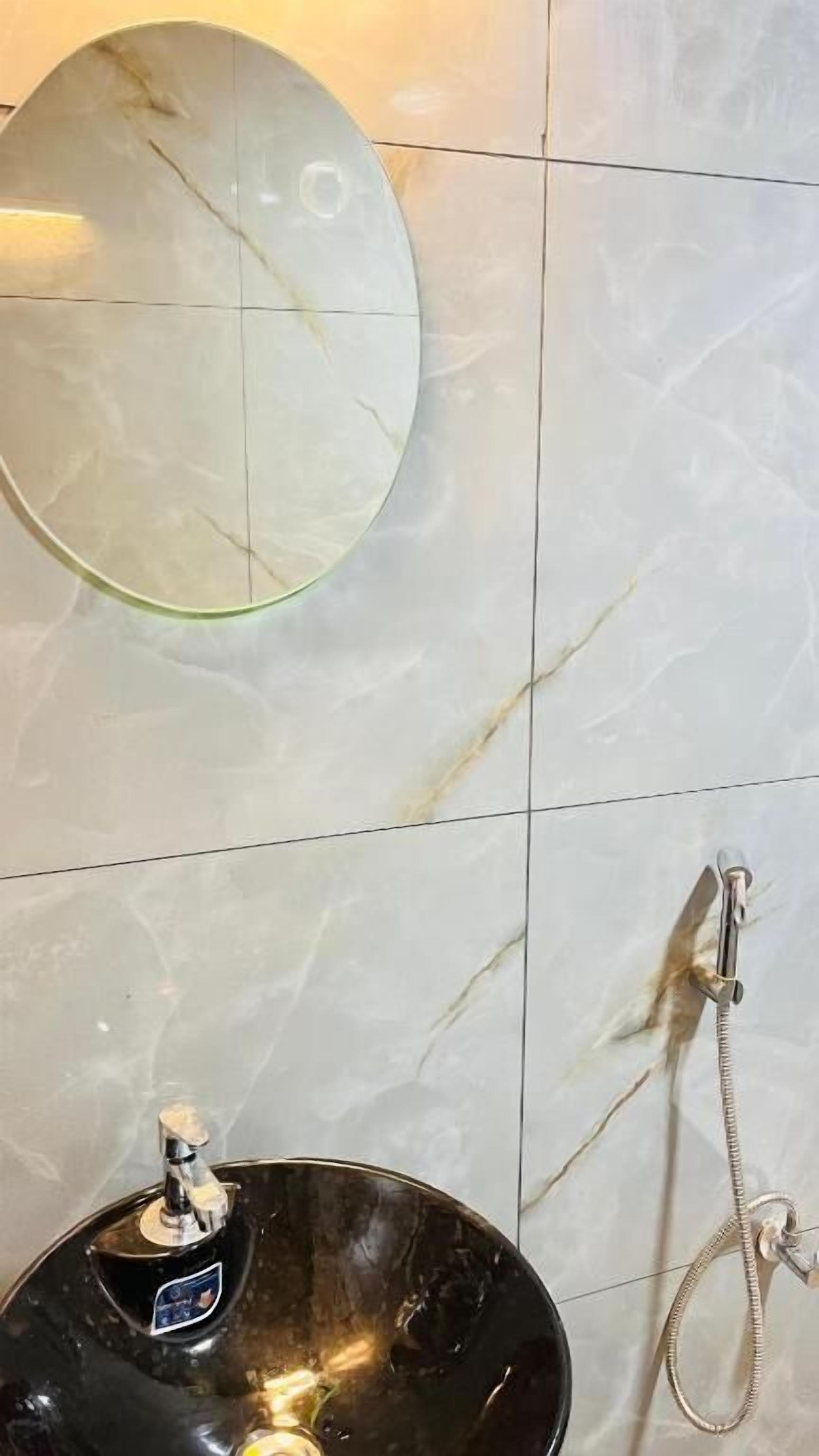 Une salle de bain avec un mur en marbre, un miroir rond, un lavabo avec un receveur noir et une douchette à main.