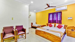 A hotel room with a large bed, two chairs, a ceiling fan, an air conditioning unit, and a window with purple curtains. - Hommetree T Nagar (Chennai)