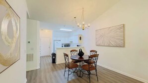 A dining area with a round table and six chairs, a chandelier, and a kitchen in the background. - Cozy 2BR condo at River Oaks (Myrtle Beach)
