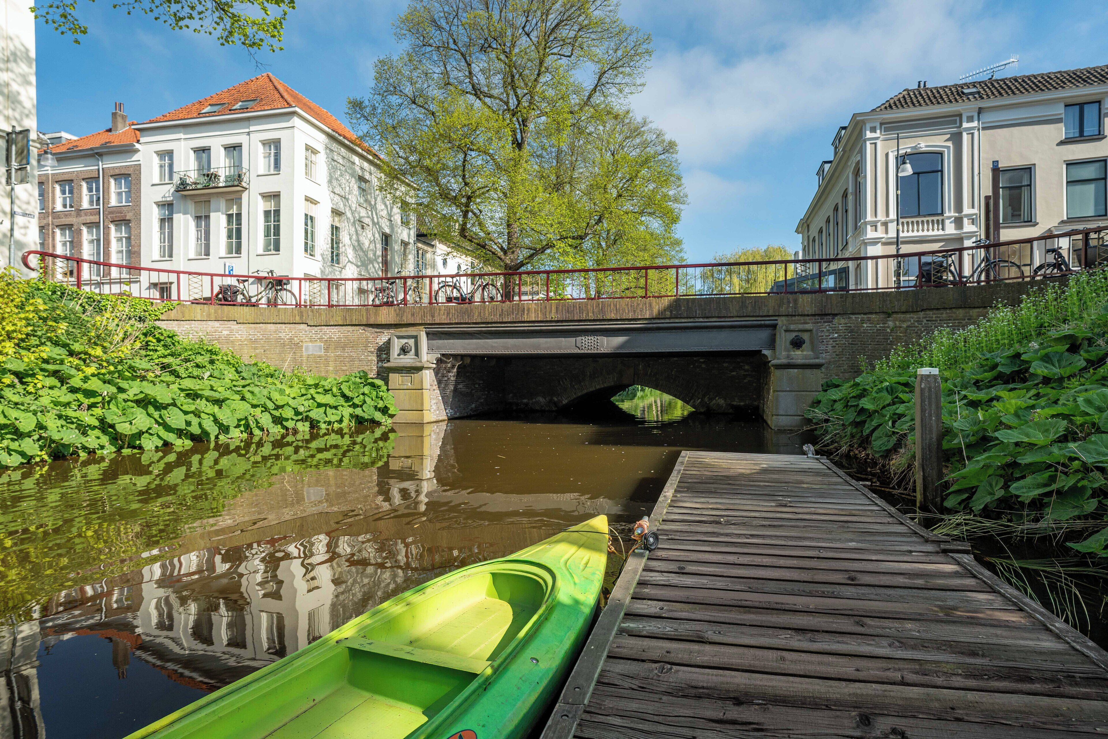 A canal with a green boat docked on a wooden pier, a bridge, and buildings with balconies.