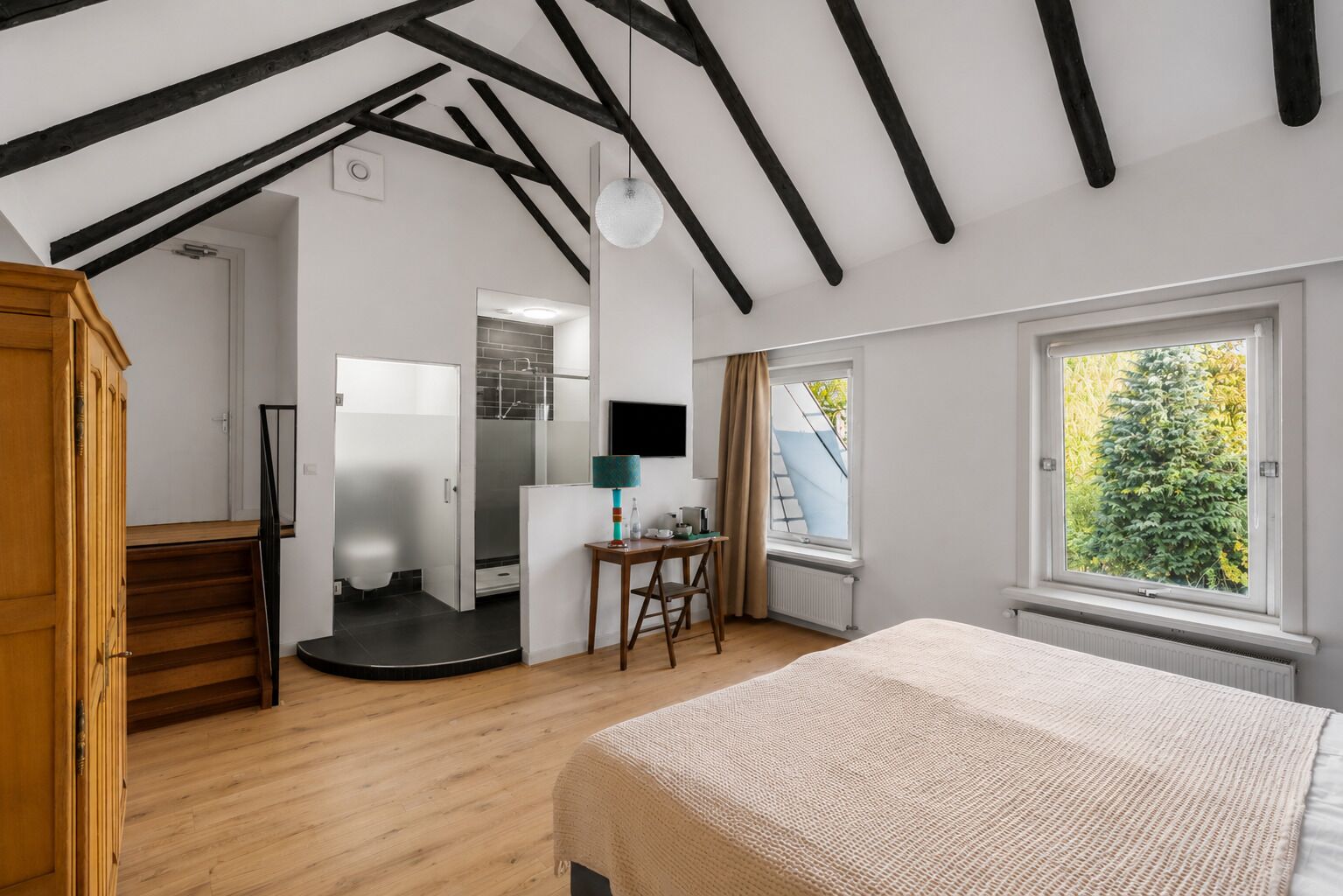 A room with a sloped ceiling, wooden staircase, a bed, a desk with a lamp, and a window with a view of trees.