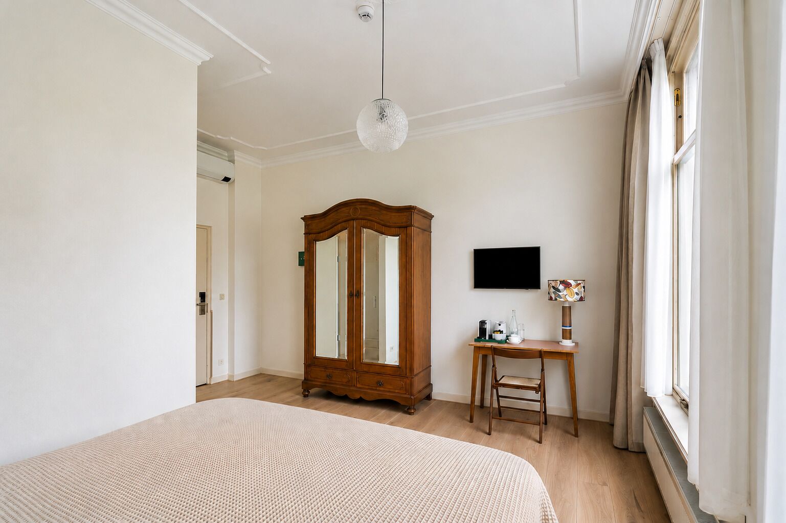A bedroom with a large bed, a wooden wardrobe, a desk with a chair, a television, and a window with curtains.