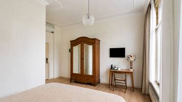 A bedroom with a large bed, a wooden wardrobe, a desk with a chair, a television, and a window with curtains.