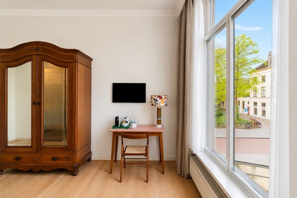 A room with a wooden cabinet, a small table with a chair, a flat-screen TV, and a window with a view of buildings and a street. - Café Hotel De Prins (Zutphen)