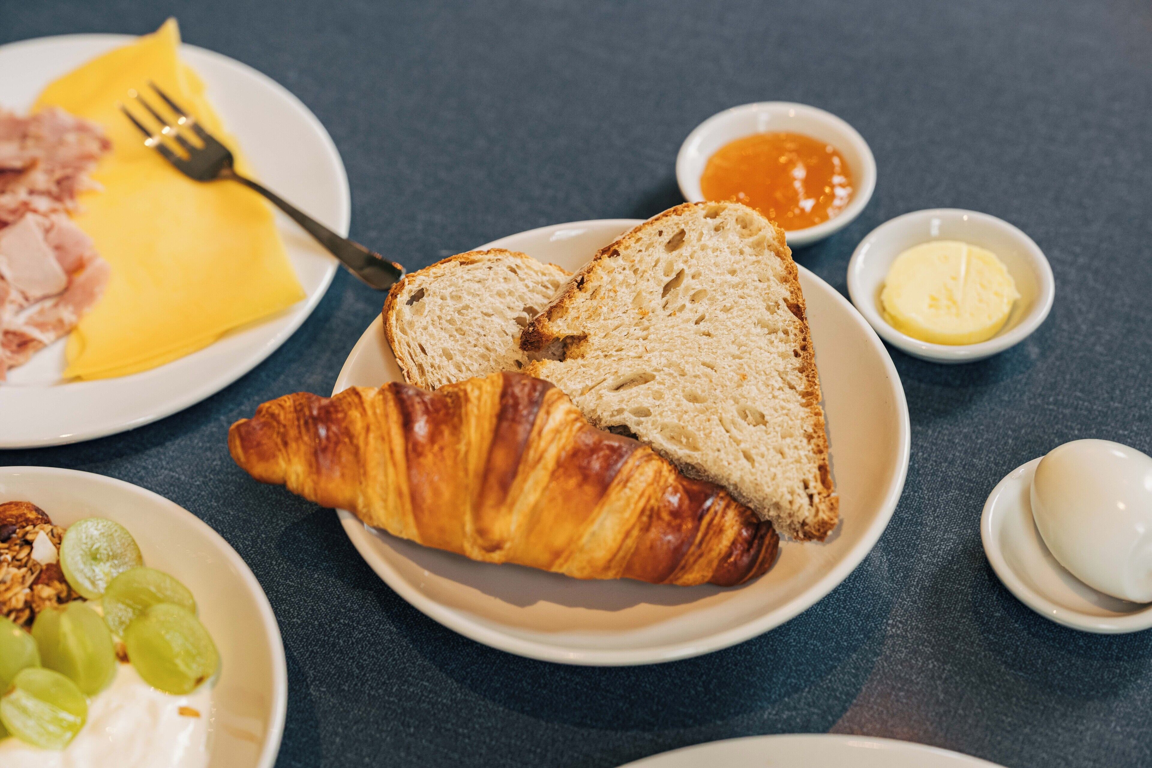 Une table dressée pour le petit-déjeuner avec des croissants, des toasts, du jambon et des fruits sur des assiettes.