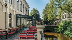 Un café al aire libre con bancos y mesas rojos, un canal y un puente al fondo.