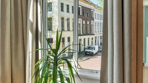 A room with a large window, a potted plant, and a view of buildings outside. - Café Hotel De Prins (Zutphen)