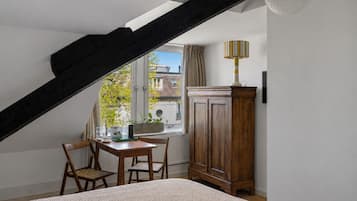 Un dormitorio con techo inclinado, un armario de madera, una mesa con sillas y una ventana con vistas a edificios.