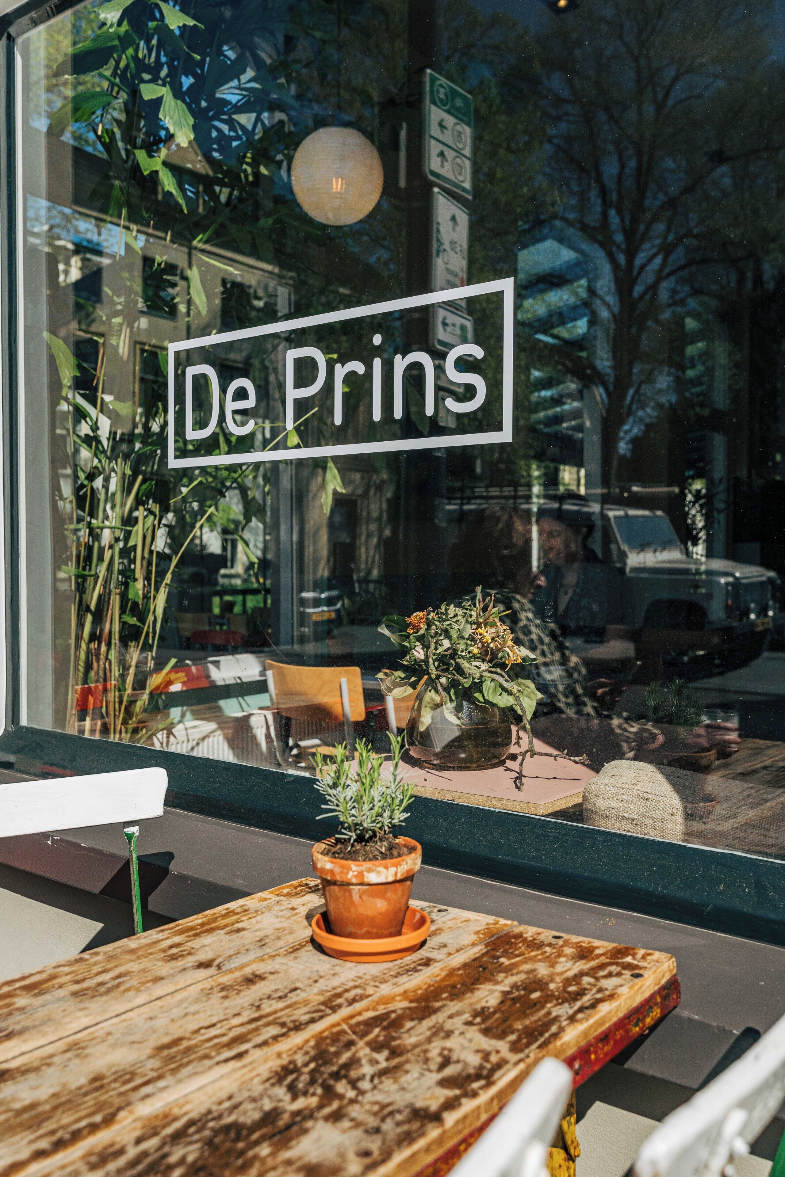 Een raam met het bord 'De Prins' en een bloempot op een houten tafel.