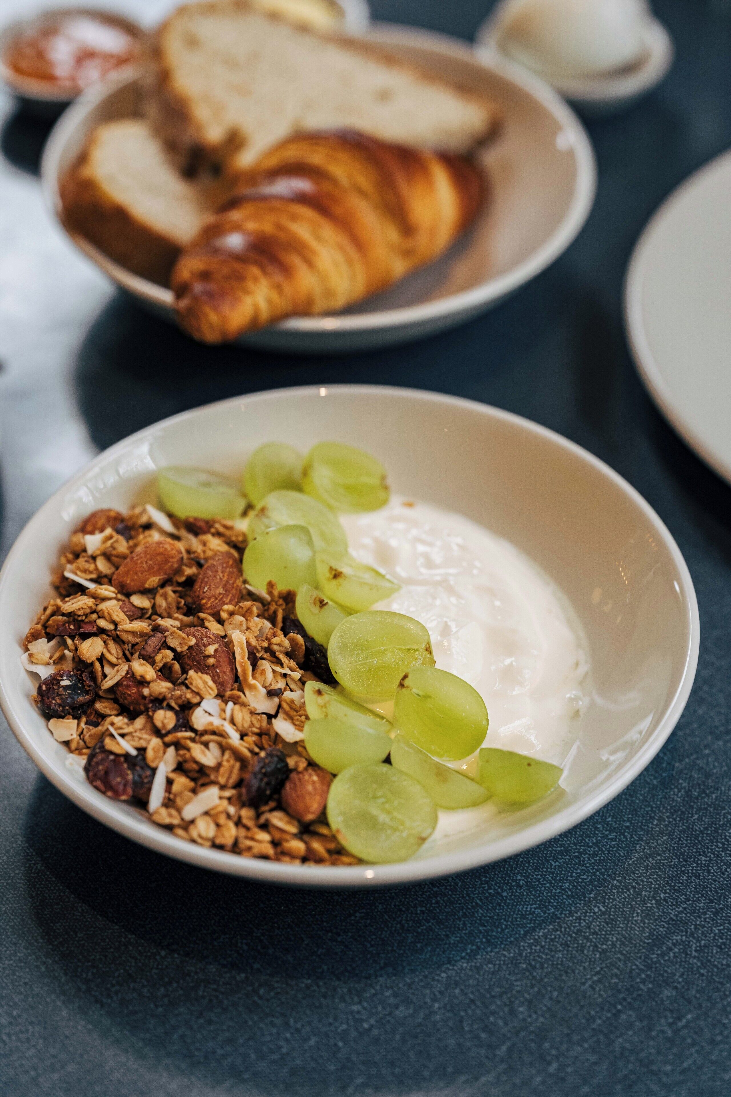 Uma tigela de iogurte com granola e uvas, um croissant e fatias de pão sobre uma mesa.