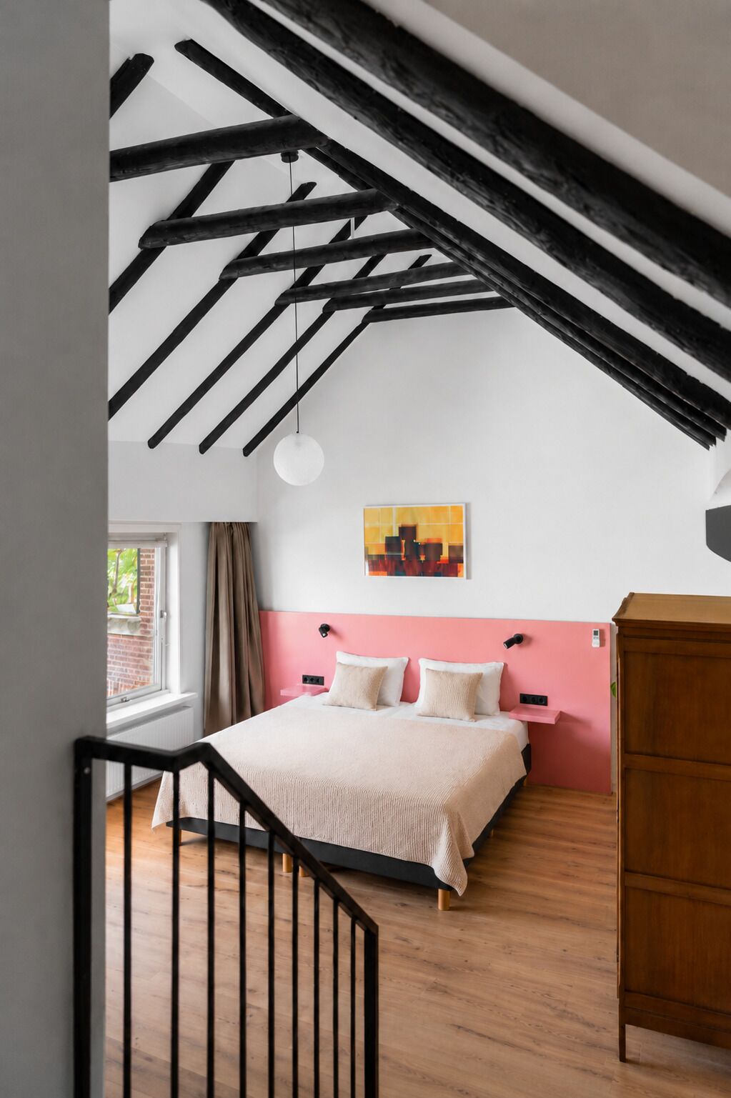 A bedroom with a sloped ceiling, a bed with white and pink bedding, a wooden dresser, and a painting on the wall.