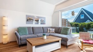 Ett modernt vardagsrum med en grå soffa, ett träbord och utsikt över ett hus genom ett stort fönster.