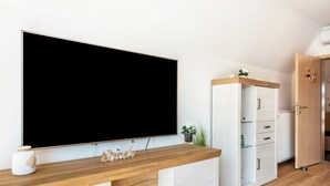 Ett modernt rum med en stor TV med platt skärm, ett skrivbord i trä, ett vitt skåp och en dörr som leder till ett annat rum.