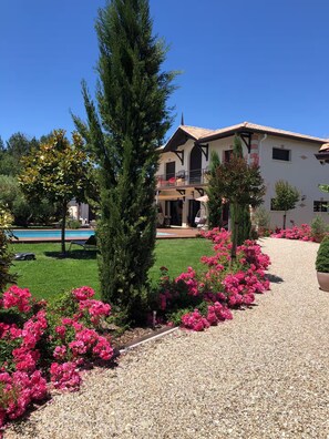 Une maison à deux étages avec une piscine, entourée d’un chemin de gravier et de fleurs roses éclatantes.