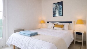 A neatly made bed with white bedding and blue towels, two bedside lamps, a painting on the wall, and a window with blinds.