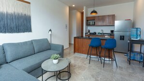 Ein modernes Wohnzimmer mit einem grauen Sofa, einem kleinen runden Tisch mit einer Pflanze und zwei blauen Barhocker. Ein Küchenbereich ist sichtbar, mit einem Kühlschrank, einer Mikrowelle und einem Wasserspender.