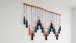 Ein ordentlich bezogenes Bett mit weißen Leinentüchern und einem türkisfarbenen Kissen, ein Nachttisch mit einer Lampe sowie ein wandmontiertes Fransenornament.
