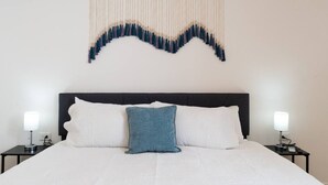 Een net opgemaakt bed met een witte deken en twee kussens, een donker hoofdbord en een decoratief object aan de muur erboven.